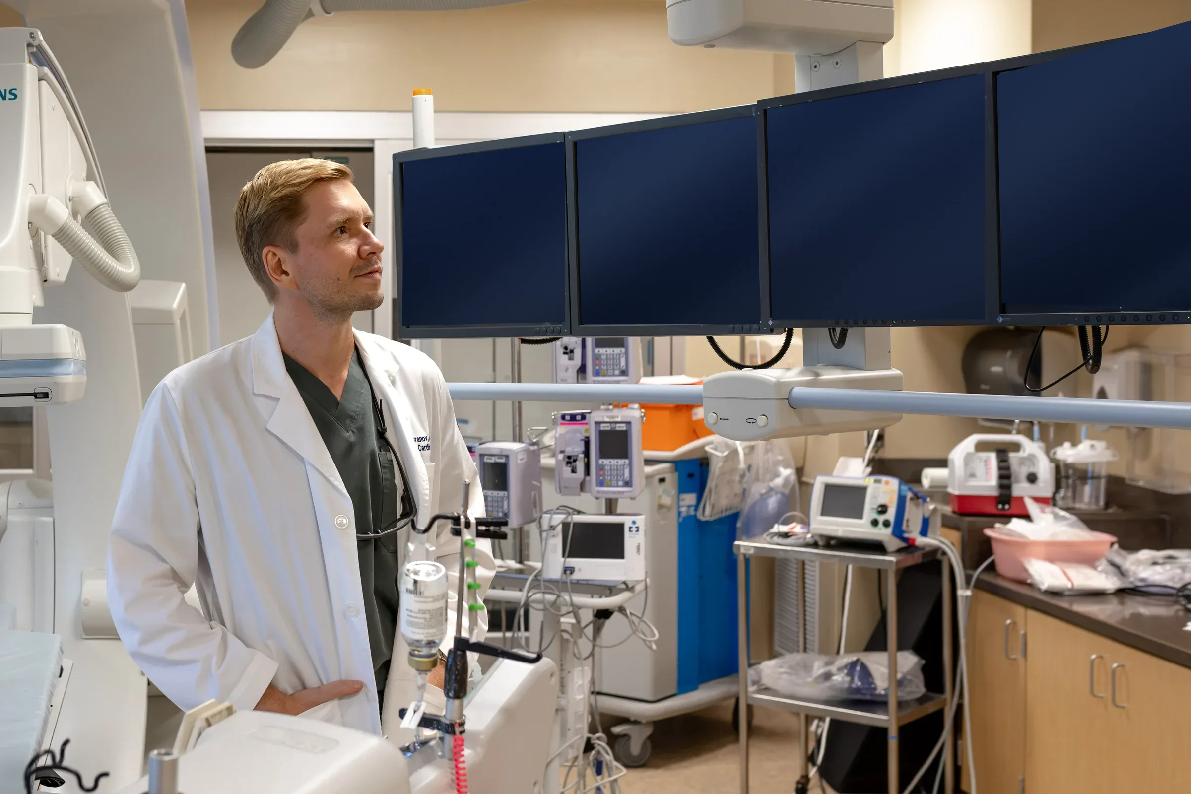 Dr. Yaranov in a white coat in the cath lab, looking up at a bank of monitors beside an angiography system