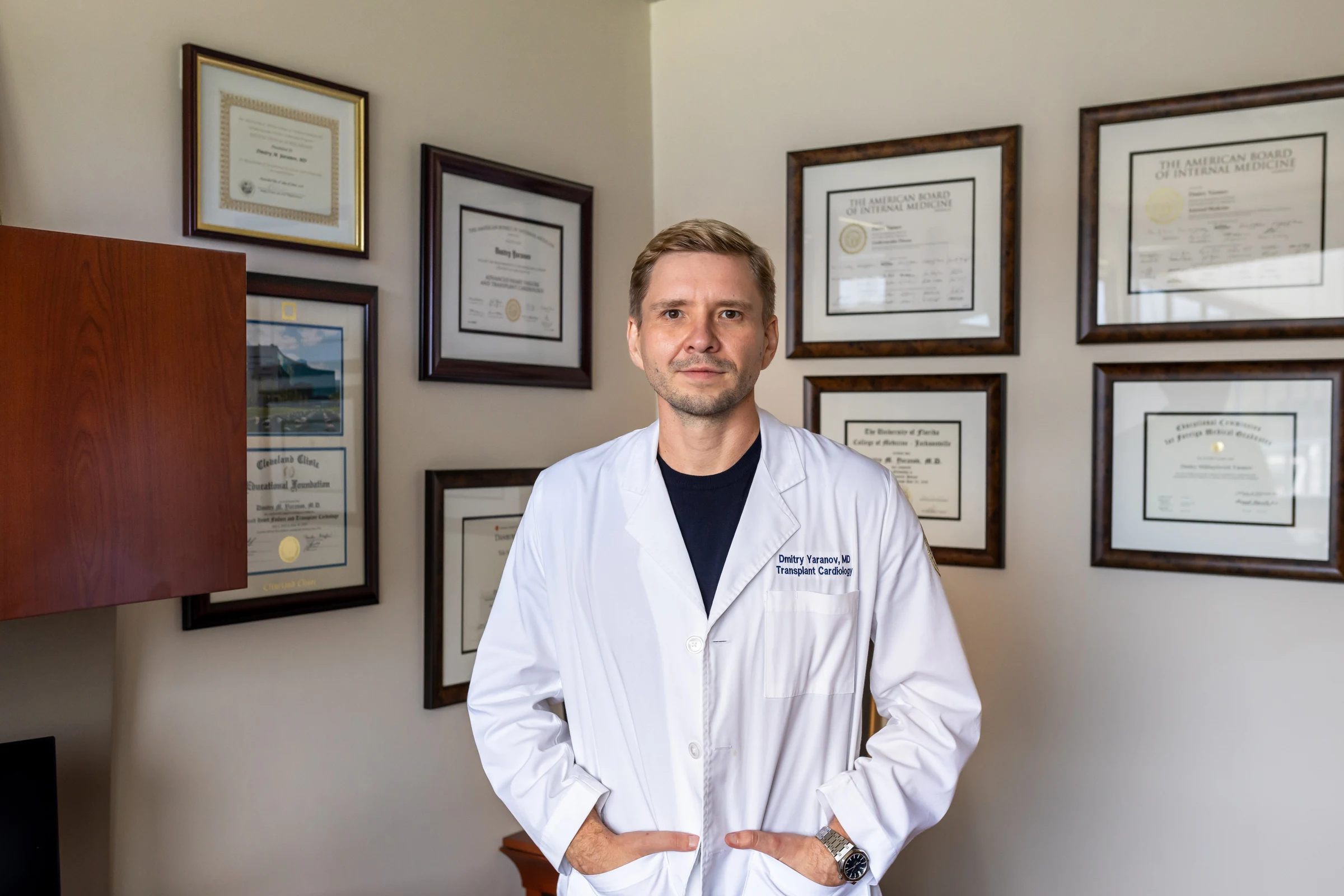 Dr. Yaranov in a white coat in front of his framed diplomas